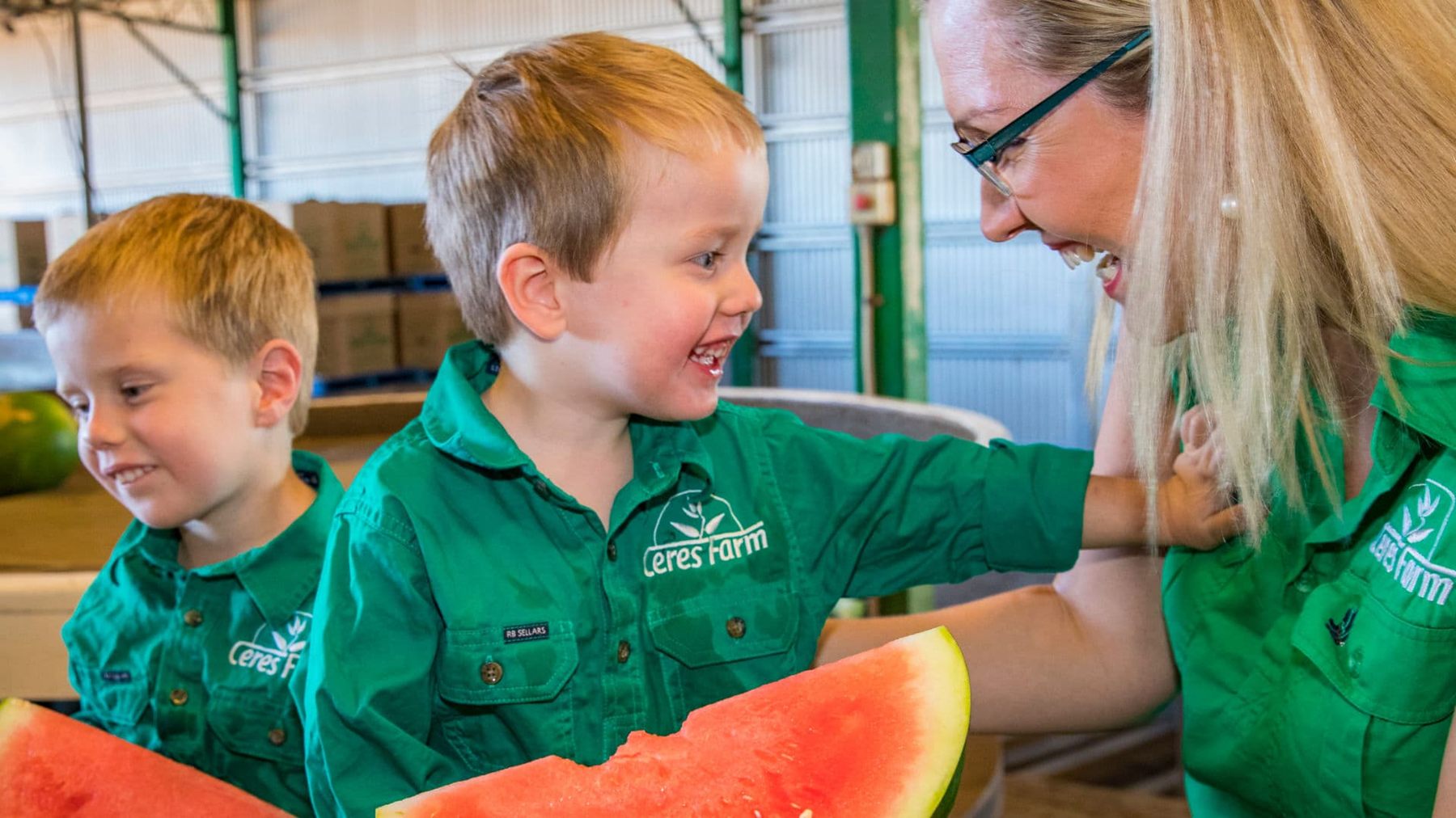 Melanie Gray laughing with her two young sons, all in green Ceres Farm shirts, holding slices of watermelon in the packing shed